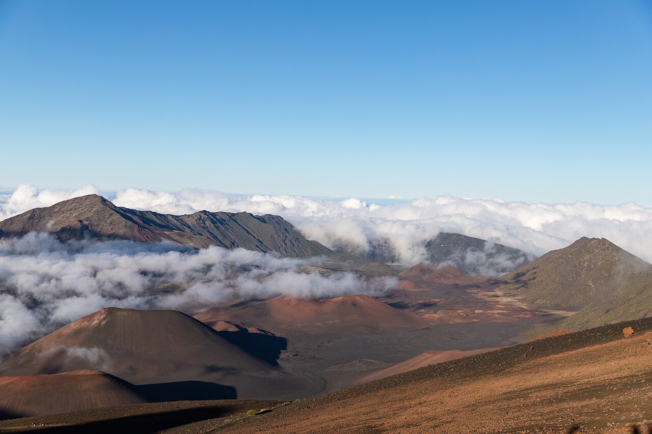 Red and brown cinder cones on the floor of Haleakalā’s summit crater on Maui, with cloud drifting below the crater rim under a clear blue sky