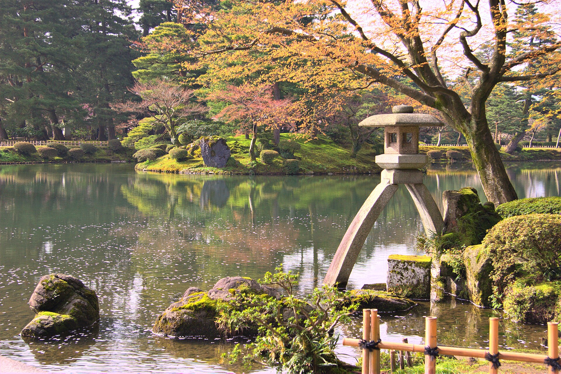 The two-legged Kotoji stone lantern standing at the edge of Kasumigaike pond in Kenroku-en, with pines and autumn-toned trees reflected in the water