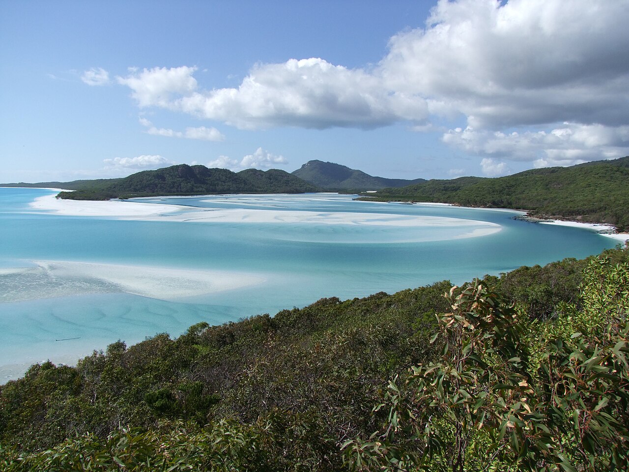 Swirls of white silica sand and turquoise shallows at Hill Inlet, seen from the lookout above, with forested hills behind